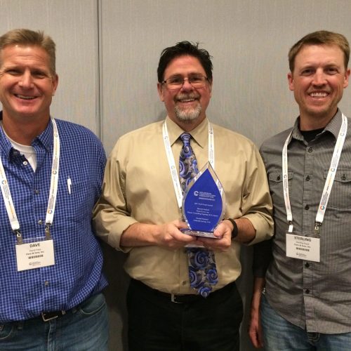 Three men are standing together; the middle man holds an award. All are smiling and wearing nametags.