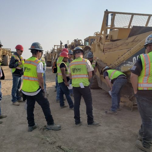 Construction workers in safety vests and helmets gathered around machinery on a dirt site, inspecting and discussing equipment.