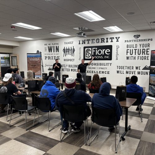 A group of people sit in a room facing two speakers. The wall displays motivational words and phrases.