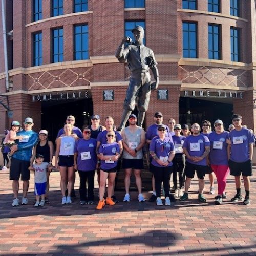 Group of people wearing purple shirts and race bibs, standing in front of a large baseball player statue and a building labeled "Home Plate.