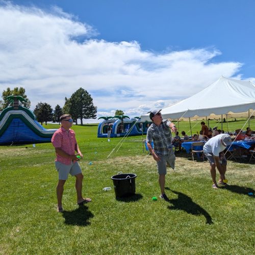 Three people stand on grass playing a game near inflatable slides and a white tent. A group is seated under the tent on a sunny day.