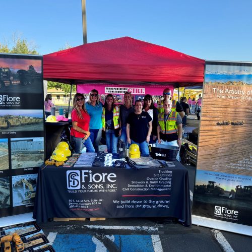 A group of people stands under a red canopy at an outdoor event, behind a table with promotional materials for Fiore & Sons, Inc. Two large informational banners flank the table.
