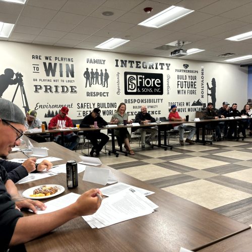 People seated around a U-shaped table in a meeting room with motivational words on the wall. One person in the foreground is eating while reading a document.