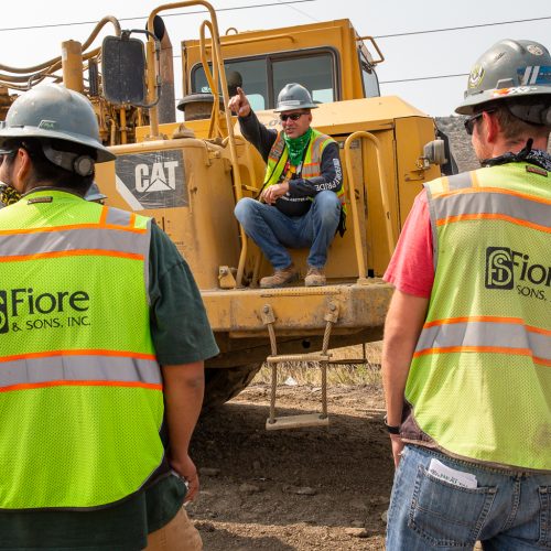 Construction workers wearing safety vests and helmets gather near heavy machinery for a briefing. One worker sits on the equipment, gesturing as others listen.