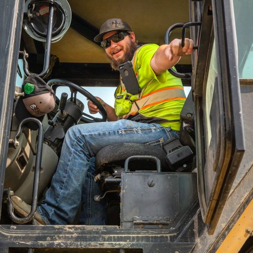 A man in a high-visibility vest and hat sits in the driver's seat of a large vehicle, smiling and holding onto the door.