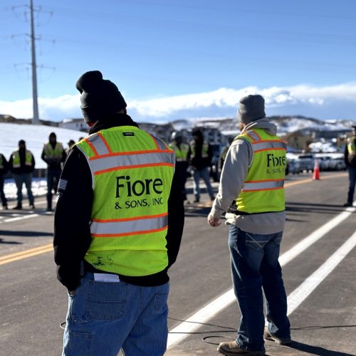 Construction workers wearing high-visibility vests with "Fiore & Sons, Inc." stand on a paved road with snow and mountains in the background.